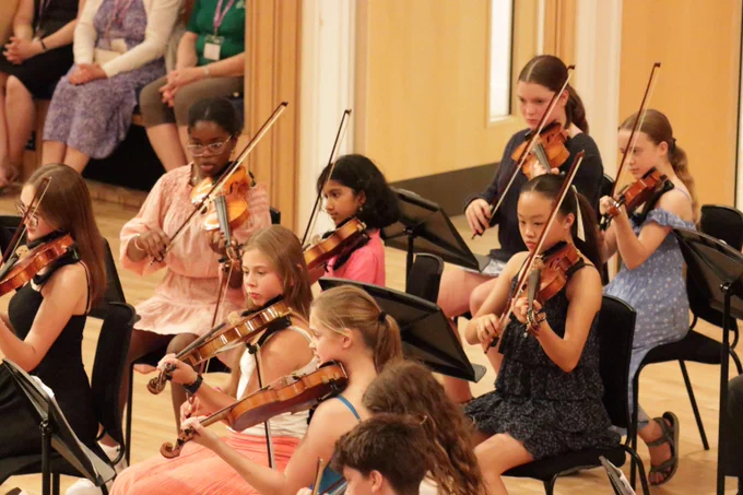 Young musicians playing violins in a seated orchestra rehearsal inside a well-lit room.
