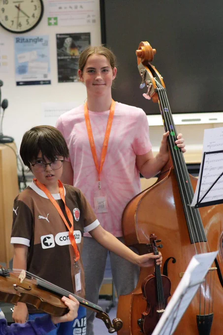Two students in a music classroom holding string instruments, one with a violin and the other with a double bass, preparing to play.