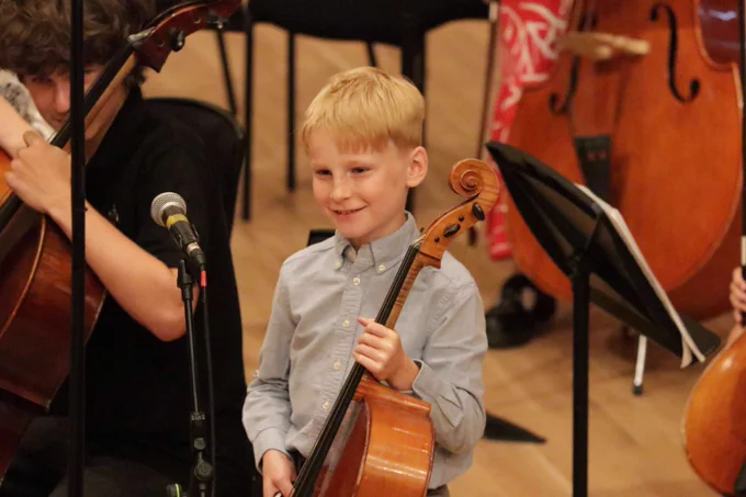 Young boy in a light blue shirt holding a cello during a music rehearsal with other string musicians nearby.