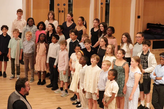 A diverse group of children standing in rows on a wooden floor, singing together in a choir setting inside a room with wooden panel walls.