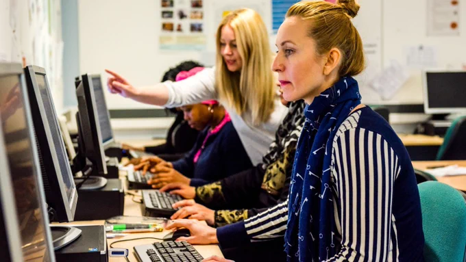 A photo of a woman sat at a desktop computer. She is looking at the screen and using the mouse. Next to her, other students are sat using computers. A tutor is leant forward and pointing to one of their screens. 