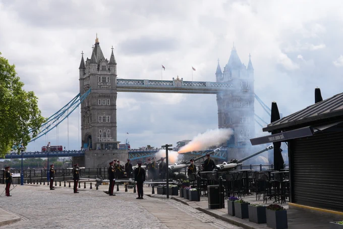 A ceremonial cannon is being fired outside the Tower of London, in front of Tower Bridge. Uniformed individuals are present, with smoke visible from the cannon blast. The iconic bridge looms in the background under a cloudy sky.