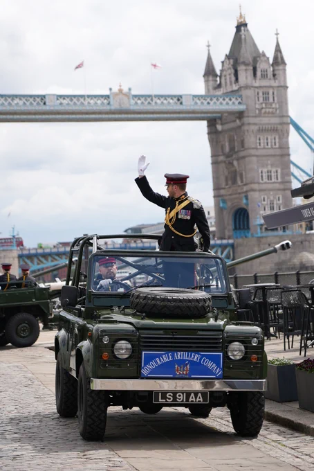 A person in a formal uniform is standing with his hand raised in an open-top military vehicle near Tower Bridge in London. The vehicle has a sign that reads "Honourable Artillery Company." The bridge and surrounding area are recognisable landmarks.