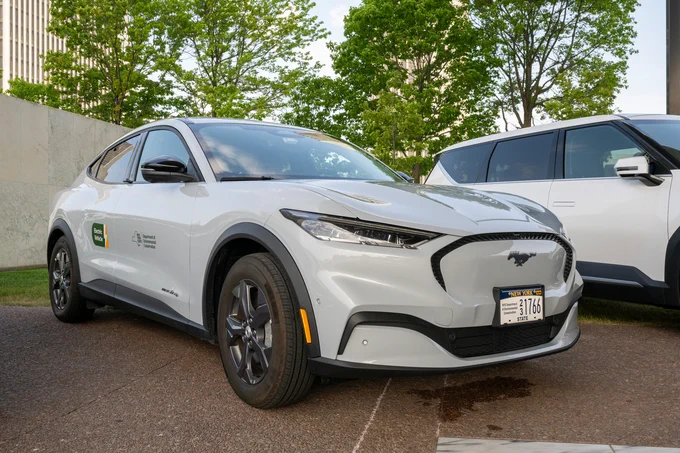 A white Ford Mustang Mach-E with New York State Electric Vehicle branding parked outdoors near trees, with another electric SUV beside it.