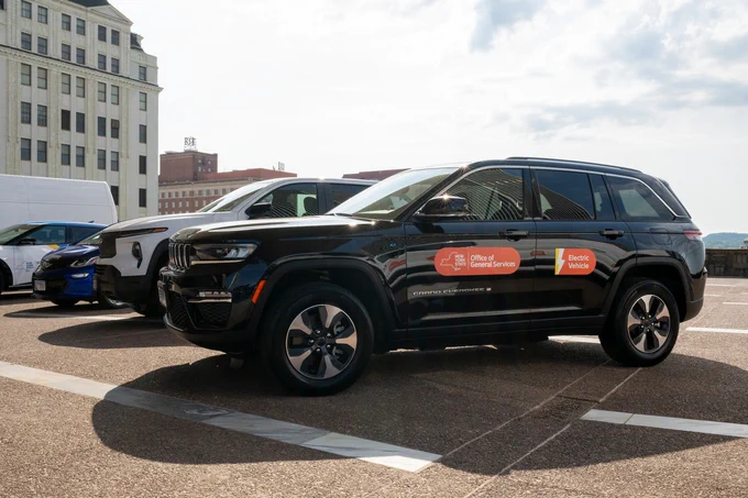 A lineup of electric SUVs, including a black Jeep Grand Cherokee 4xe with New York State Office of General Services branding, parked in an urban lot with city buildings in the background.