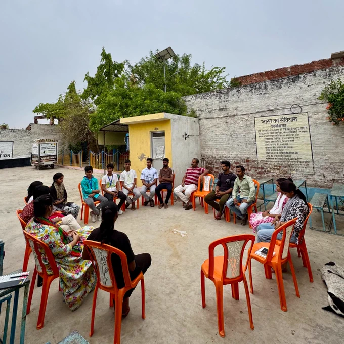 A group of young people and adults sit in a circle on orange plastic chairs in an open courtyard, engaged in a discussion. The setting appears to be a village space with boundary walls, trees, and a small structure in the background. Some participants are listening attentively while others are speaking, creating a sense of an open, inclusive conversation. A dog lies nearby, and the overall atmosphere reflects a calm, safe community space for dialogue.
