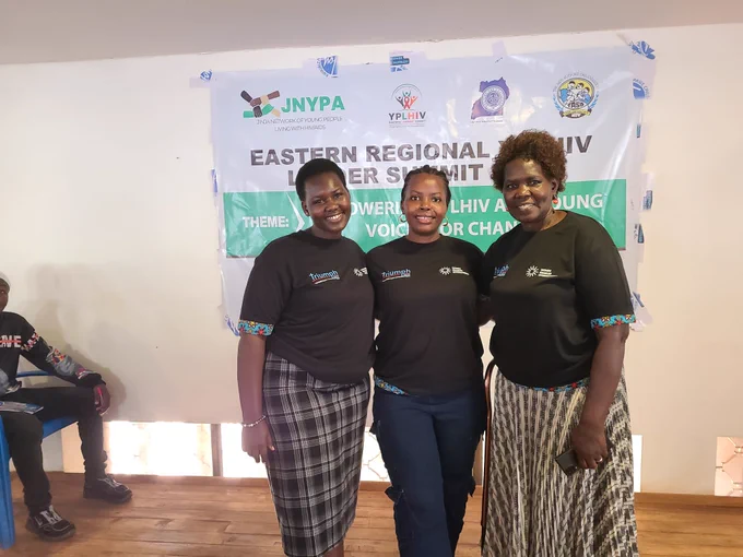 A medium shot of three women standing together and smiling in front of the summit banner. They are all wearing matching black TRIUMPH t-shirts, and the woman on the right is resting her hand on a mobility cane.