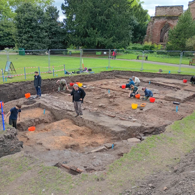A view of the excavation trench surrounded by green grass, the ruined east end of the Church of St John the Baptist is in the background. The orange Roman ground surface is in the foreground, the foundations of walls and a cobbled surface are behind.