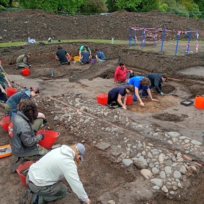 Students are trowelling in the excavation trench , they have brightly coloured buckets and are dressed in blue, white, red and grey.