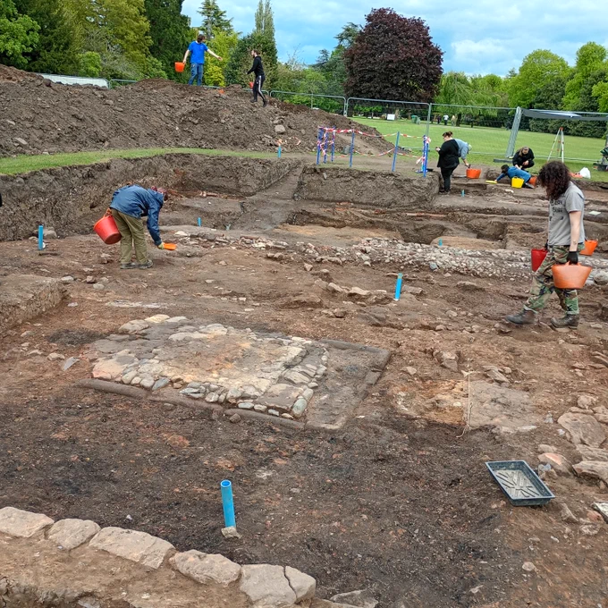 A view of the excavation trench looking eastwards, grass and trees are beyond the trench.  in the foreground is a line of pale stone blocks making a wall,  An area of dark soil separates the wall from a square oven base or hearth. Students are carrying orange buckets