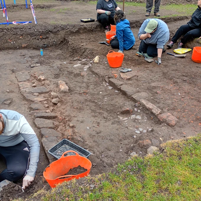Students dressed in blue and grey pullovers, they are trowelling and putting the soil into orange buckets. In the centre of the trench are two parallels lines of stone blocks.