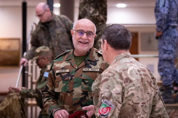 Two military personnel from different countries engaged in conversation indoors, one wearing Pakistan Navy uniform and the other in camouflage with a British flag patch.