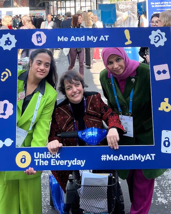 Three women pose together at an event behind a large blue ‘Me And My AT’ photo frame from the Unlock The Everyday campaign. One woman is seated on a mobility scooter, while the frame is decorated with icons representing assistive technology such as hearing aids, glasses, wheelchairs, prosthetics, and digital devices. The hashtag #MeAndMyAT appears at the bottom.