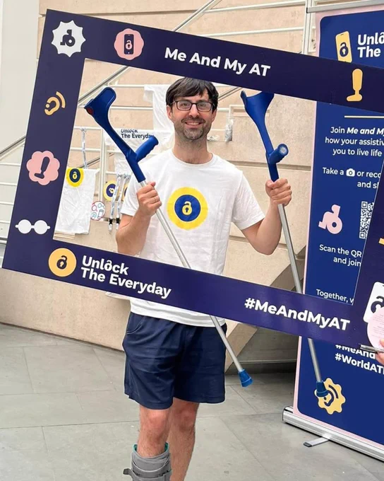 Man standing indoors and smiling while holding a pair of blue crutches behind a large ‘Me And My AT’ campaign photo frame from Unlock The Everyday. He wears a white campaign T shirt and a medical walking boot on one leg. The frame includes icons representing assistive technology and the hashtag #MeAndMyAT.