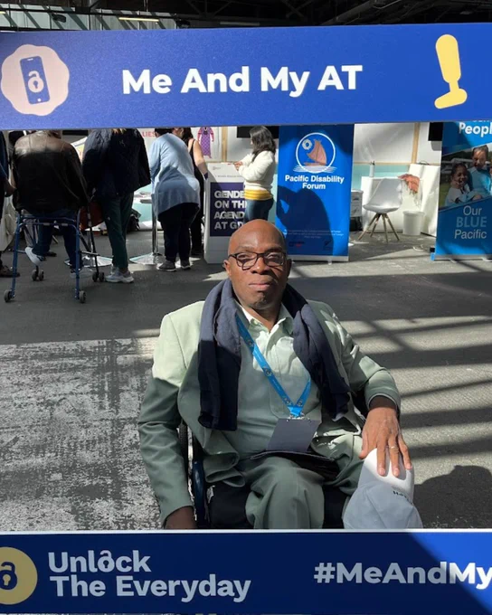 Man seated in a wheelchair at an event behind a large blue ‘Me And My AT’ campaign photo frame from Unlock The Everyday. The frame features assistive technology icons and the hashtag #MeAndMyAT, with exhibition booths and attendees visible in the background.