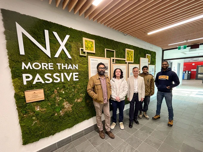 A group of people are standing together next to a wall that reads NX More Than Passive.
