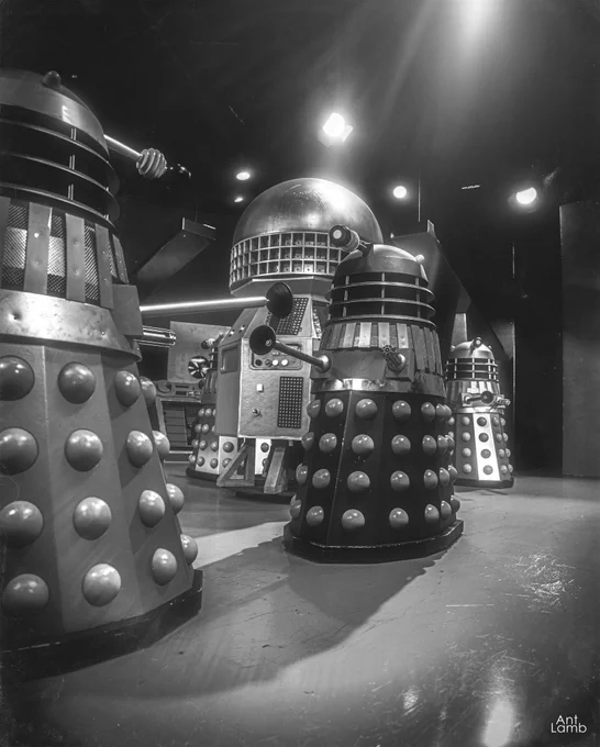 A black and white CG render of the Dalek control room. The black Supreme Dalek is in the middle, with out of focus Silver Daleks in the foreground and background. A large central control unit sits behind the Supreme.