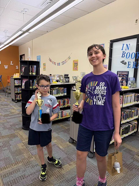 Two children proudly hold trophies in a library setting.