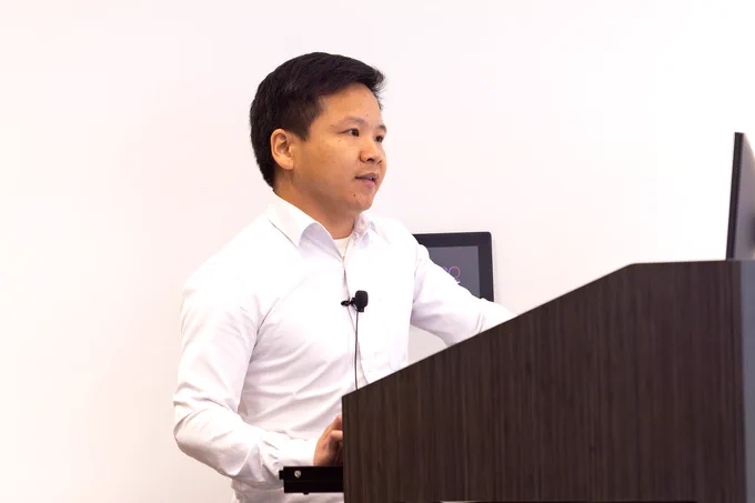 A man stands behind a podium and gives a presentation on his scientific research to a room full of researchers and students.