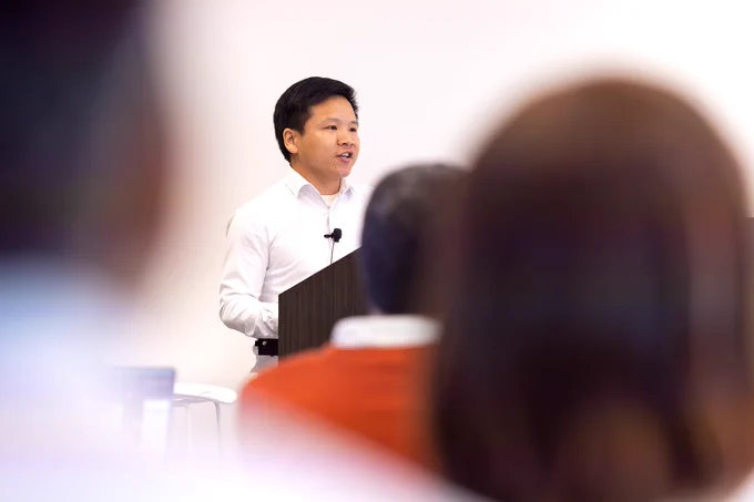 A man stands behind a podium and gives a presentation on his scientific research to a room full of researchers and students.
