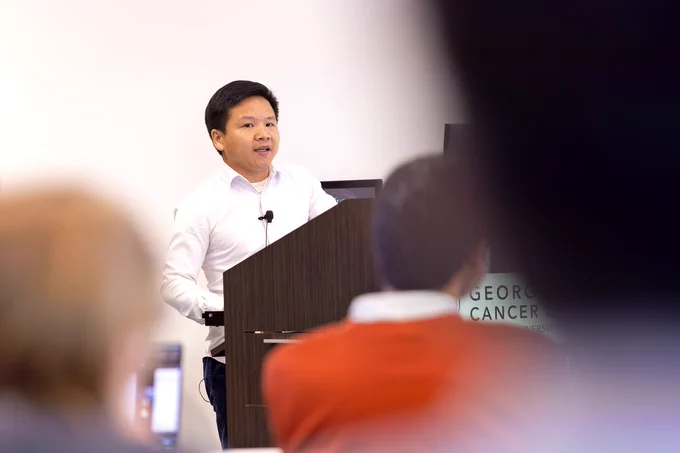 A man stands behind a podium and gives a presentation on his scientific research to a room full of researchers and students.