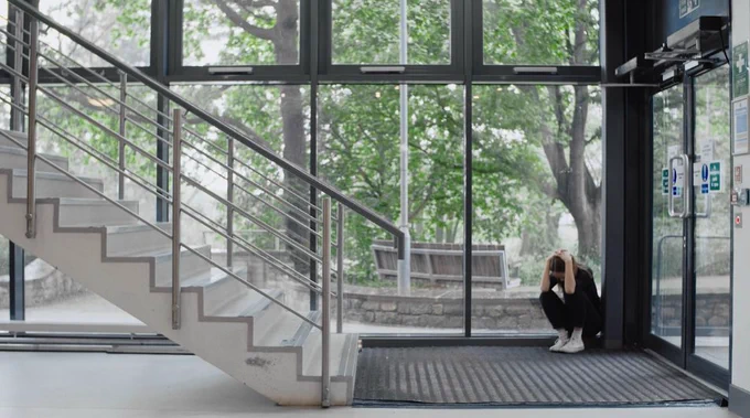 A person is sitting on the floor near a wall of large glass windows with visible trees outside. They are holding their head in their hands, seated near an ATM and a staircase.