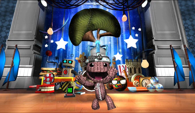 Sackboy smiles for the camera as he wears a sci fi crown. Wires power a PlayStation move controller on the top of the crown. The background contains a mess of creative tools, bits and bobbles, and the iconic LBP mushroom tree against a backdrop of curtains, flags, and ornate pillars.