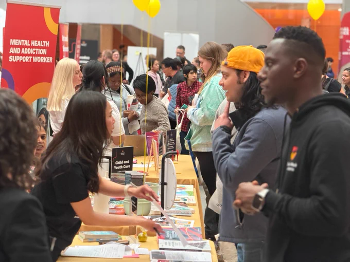 A bustling open house with many attendees interacting at various booths. One booth features a sign reading "Child and Youth Care." Another sign mentions "Mental Health and Addiction Support Worker." People are engaged in conversations, and informational materials are displayed on tables. Bright balloons are visible in the background.