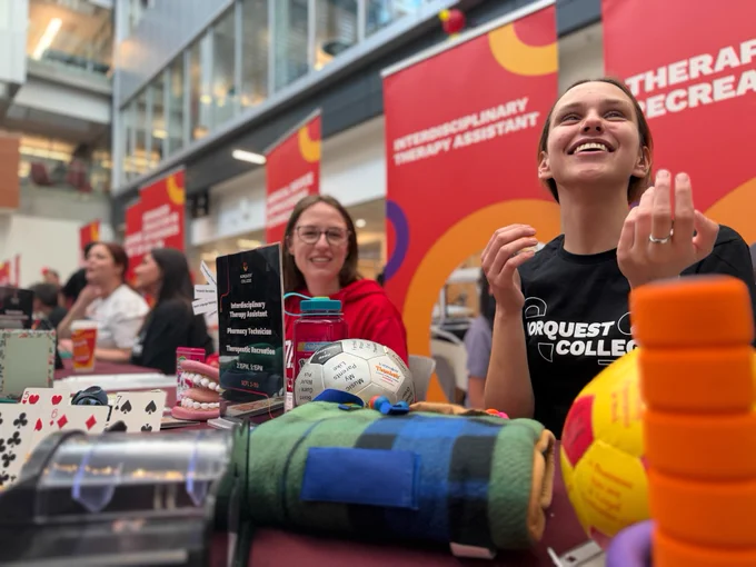 A lively indoor event at NorQuest College features tables displaying various therapy and recreational items. Several individuals are seated behind the tables, engaged in conversation. Prominent red and orange banners in the background read "Interdisciplinary Therapy Assistant" and "Therapy and Recreation."