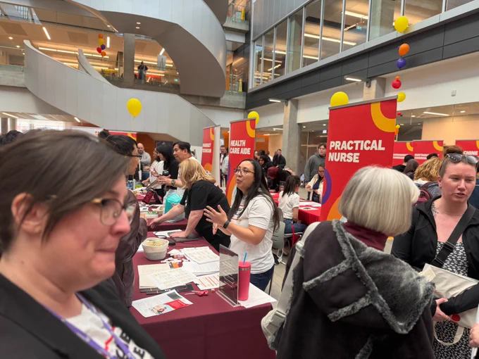 A busy indoor career fair with people interacting at various booths. Visible signs include "Health Care Aide" and "Practical Nurse." Bright yellow and red balloons are floating above. A multi-level staircase and glass railings are in the background.