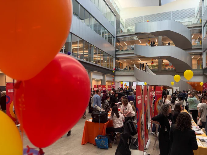 A bustling open house in a spacious atrium with multiple floors visible. Booths staffed by people are lined up, with prominent signs for various programs. Bright balloons in red, orange, and yellow are decorating the space, and a spiral staircase in the background connects the different levels. Attendees are engaging and exploring the various booths.