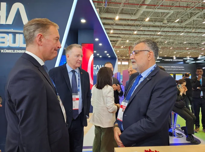 Two men in suits stand and talk with another man in a suit among the booths of a trade exposition 