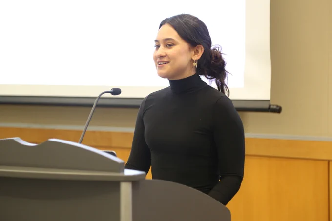 Woman in a black turtleneck speaking at a podium with a microphone in a conference room.