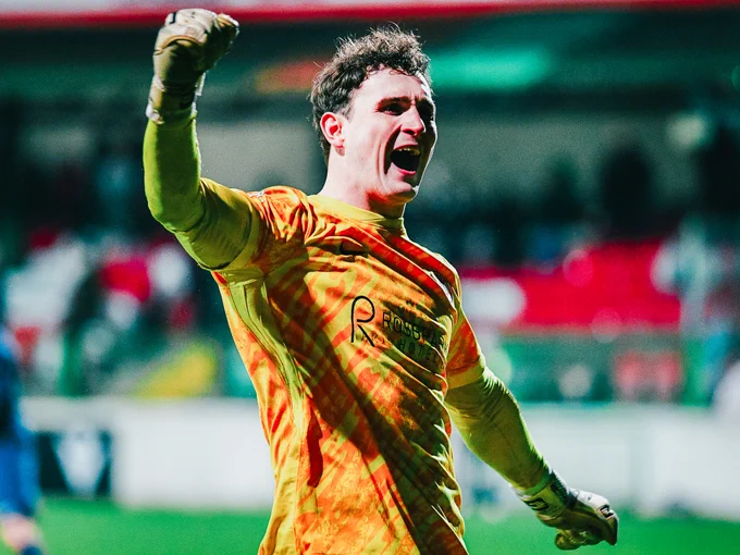 An enthusiastic goalkeeper in a yellow jersey raises his fist in celebration during a soccer match. The image captures a moment of triumph and excitement on the field.