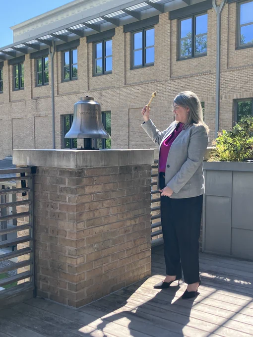 A person in a gray blazer stands outdoors beside a large metal bell mounted on a brick pedestal, holding a small mallet raised as if about to ring it. The scene is set on a deck outside a brick campus building with tall windows and metal awnings overhead. Sunlight casts strong shadows across the space. Faint text is engraved on the bell, though it is not fully legible in the image.