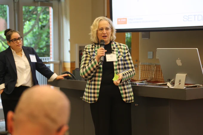Woman in a checkered blazer speaking into a microphone during a presentation in a conference room.