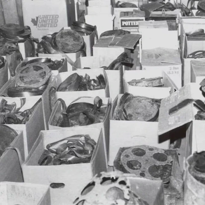 Black-and-white photo of numerous cardboard boxes arranged on the floor indoors, each filled with assorted metal objects. The items include circular plates, rings, curved fragments, and irregular pieces, many showing wear or corrosion. Some boxes are open with contents visible, while others are partially covered with paper or lids. Printed labels and markings appear on a few boxes in the background.
