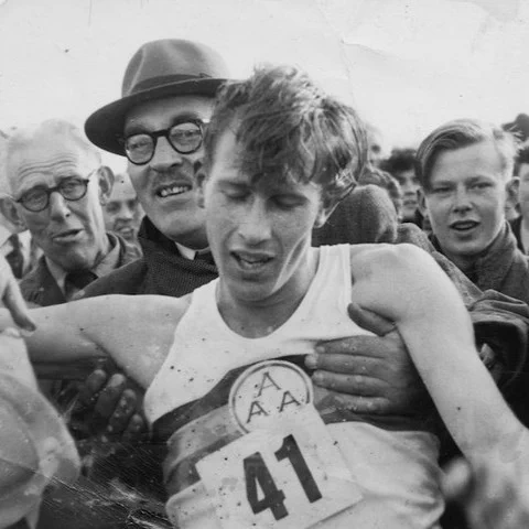 A black and white photograph of Sir Roger Bannister being supported by onlookers immediately after breaking the four-minute mile in 1954.