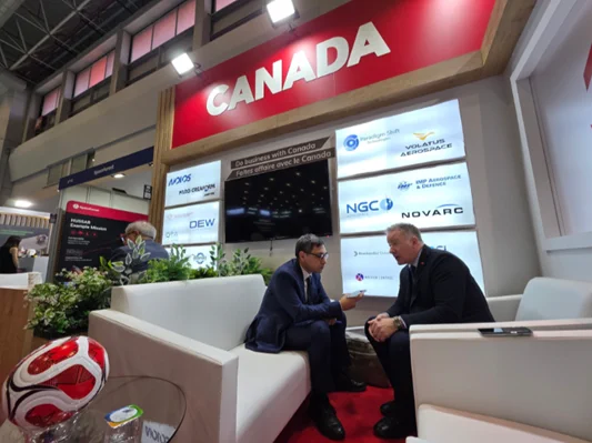 Two men sit on couches talking during a trade exhibition with booths in the background 