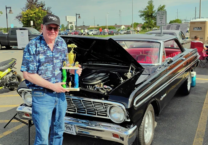 A person stands next to a classic black Ford Falcon with the hood open, holding a trophy. The car is parked in a lot with other vehicles and a motorcycle nearby.