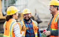 Three construction workers in safety vests and helmets are smiling and conversing at a construction site. A bulldozer is visible in the background.