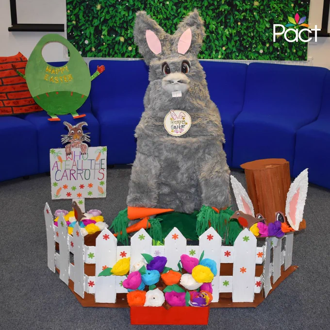 Large grey Easter bunny display surrounded by colourful paper flowers, carrots and a white picket fence, with handmade signs reading “Happy Easter” and “Hoppy Easter.”