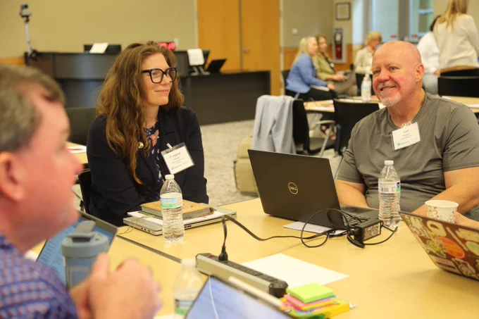 Three professionals engaged in a discussion around a table with laptops and water bottles in a conference room.