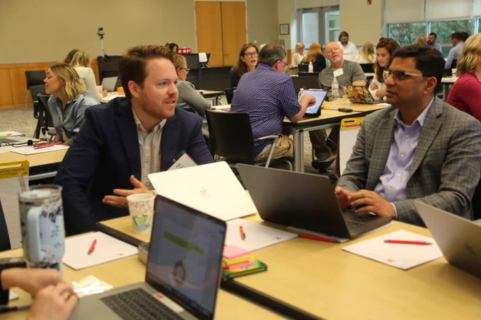 Two professionals in business attire engage in discussion at a conference table with laptops and notebooks open.