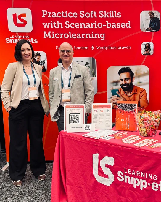 Two people stand at a "Learning Snippets" trade show booth. The red backdrop promotes "Scenario-based Microlearning" for soft skills alongside photos of phone users. A table displays brochures, a QR code, and candy.