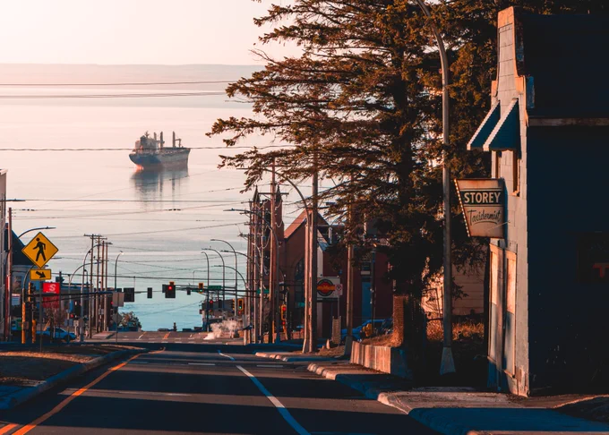 The Lake St. Clair bulk freighter is anchored in Lake Superior directly in line with Sixth Avenue. The ship is visible at the terminating view of the street. A taxidermy shop, evergreen trees, and other buildings frame the view.