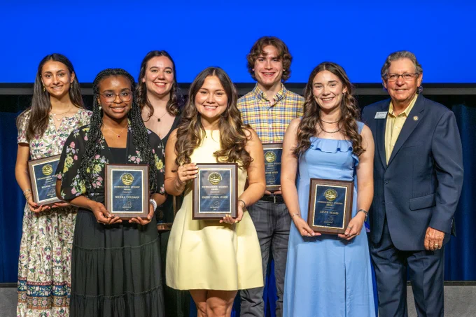Seven people stand in a row on stage, with six of them holding plaques inscribed with "DISTINGUISHED STUDENT."