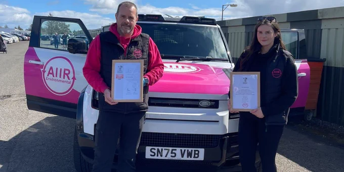 A person holding an Employer Recognition Scheme Bronze Award certificate, and a person holding an Armed Forces Covenant certificate in front of an AIIR Environmental car.