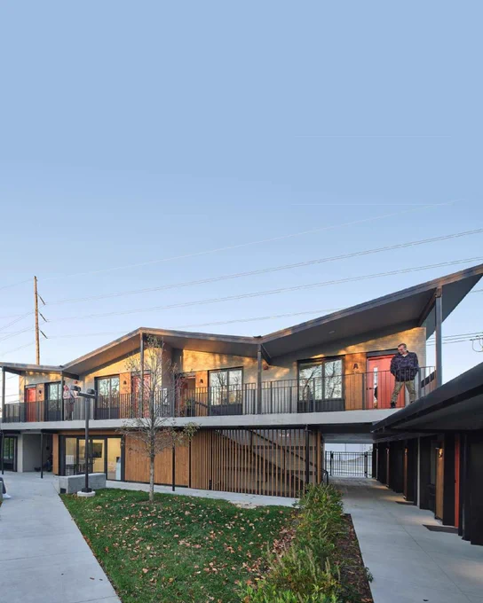 Modern two-story apartment courtyard with sloped roofs, red doors, walkways, and a few people standing on upper balconies.