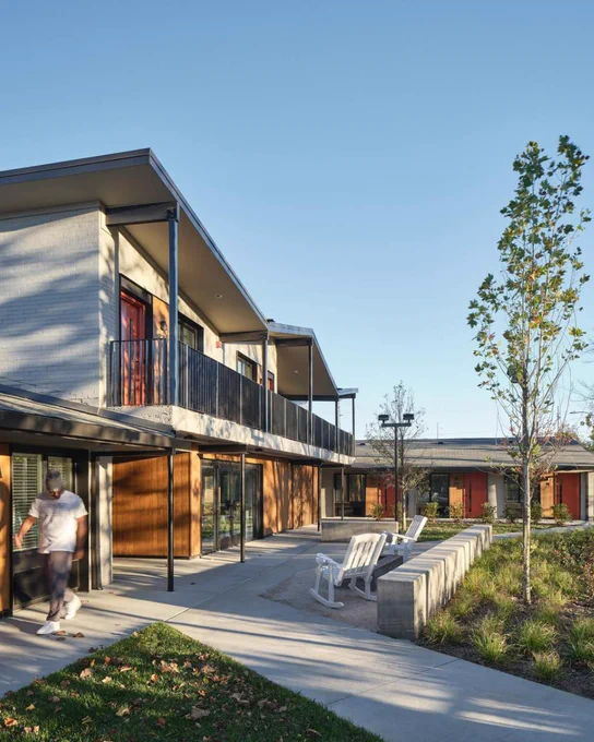 Modern apartment courtyard with two-story balconies, white rocking chairs, a young tree, and a person walking in late-afternoon light.